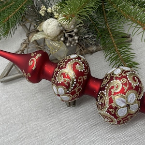 Könnte beinhalten: Ein roter Weihnachtsbaumaufsatz mit goldenen und weißen floralen Designs. Der Glasschmuck hat eine lange, spitze Spitze und zwei abgerundete Abschnitte. Das Design umfasst goldene Wirbel und weiße Blumenformen.