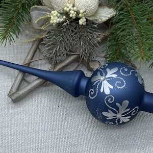 Op de afbeelding: Een marineblauwe kerstboomtop met zilveren glitteraccenten. De glazen ornamenten hebben zilveren bloemmotieven. De top heeft een lange, puntige steel en een bolvormig lichaam. Feestelijke decoratie.