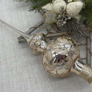 以下が含まれることがあります： 金色のグリッターアクセントが付いたシルバーのクリスマスツリートッパー。ガラス製のオーナメントは、精巧なデザインと長い尖った先端が特徴です。トッパーのベースも金色のグリッターで装飾されています。お祝いのホリデーデコレーション。