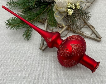 May include: A red Christmas tree topper with a long, pointed tip and a bulbous base. The base is decorated with glittery, fern-like patterns. The topper is set against a backdrop of greenery and a star-shaped decoration.