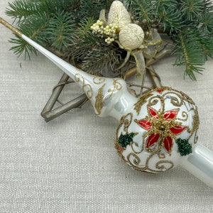 May include: A white Christmas tree topper with gold and red detailing. The topper has a poinsettia design with gold filigree and green beaded accents. The spire is white with gold glitter at the tip. A festive holiday decoration.