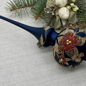 Könnte beinhalten: Ein marineblauer Weihnachtsbaumaufsatz mit goldenen Glitzerakzenten und roten, perlenbesetzten Blumendesigns. Der Aufsatz hat einen langen Stiel und eine runde, dekorierte Kugel. Festliche Weihnachtsdekoration.