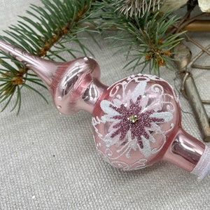 May include: A pink glass Christmas tree topper with a floral design. The topper has a long, pointed tip and a round base. The floral design is white and pink with glitter accents. The base is trimmed with white glitter.