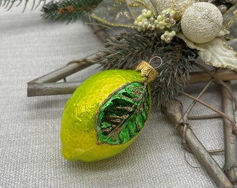 Op de afbeelding: Een kerstversiering in de vorm van een limoen met een groen blad en gouden accenten. De versiering heeft een helder geelgroene kleur en een gouden dop om op te hangen. Onderdeel van een vakantiedecoratie.