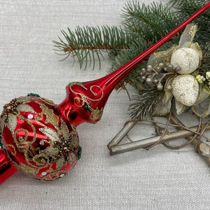 May include: A red Christmas tree topper with gold and green embellishments. The topper has a long, pointed spire and a round base decorated with gold leaf patterns and small jewels. The base is trimmed with gold glitter.