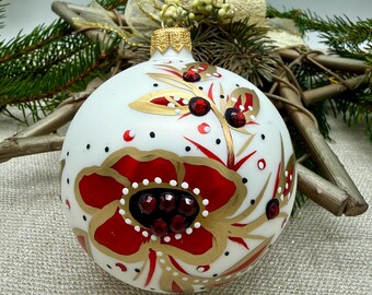 May include: White Christmas ornament with a red and gold floral design. The ornament features a large red flower with gold accents and small red jewels. Black dots and red accents are scattered across the surface. A gold cap and loop are at the top.