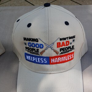 May include: A beige baseball cap with the text "Making Good People Helpless" and "Won't Make Bad People Harmless" in blue and red. Two crossed rifles are embroidered on the cap.