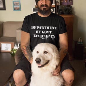 May include: Black t-shirt with the text "DEPARTMENT OF GOVT. EFFICIENCY" in white. A large white dog is in the foreground. The dog has brown eyes and a fluffy coat. The shirt also has the letters "D.O.G.E" and the pi symbol.