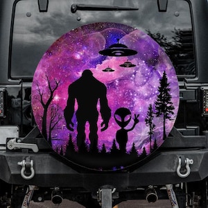May include: Black Jeep with a spare tire cover featuring a silhouette of Bigfoot and an alien in front of a purple and pink galaxy background. Three UFOs are above them. The scene is set in a forest with trees and mountains.