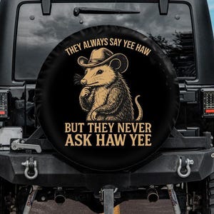 May include: Black spare tire cover featuring a cartoon opossum wearing a cowboy hat. The text reads "THEY ALWAYS SAY YEE HAW BUT THEY NEVER ASK HAW YEE" in a circular design. The cover is mounted on a black Jeep.