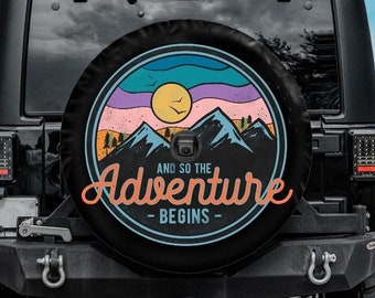 May include: Black Jeep SUV with a spare tire cover featuring a colorful mountain and sun design. The cover has the text "AND SO THE ADVENTURE BEGINS" in orange and white. The vehicle is parked on a paved road with yellow lines.