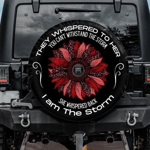 May include: A black spare tire cover with a red and pink sunflower design. The text on the cover reads "They whispered to her you can't withstand the storm. She whispered back I am the storm."