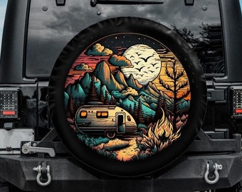 Pode incluir: Um Jeep preto com uma capa de pneu sobressalente que apresenta uma ilustração colorida de uma caravana estacionada em um ambiente de floresta com uma fogueira, montanhas e uma lua cheia no céu.
