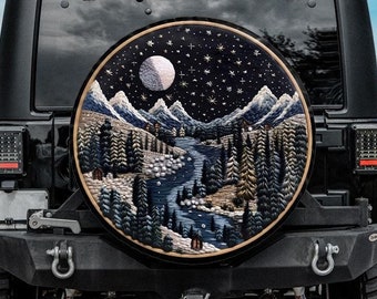 May include: A black Jeep with a spare tire that has a custom cover featuring an embroidered design of a wintery landscape with a river, mountains, and a full moon. The design is in shades of blue, white, and brown.