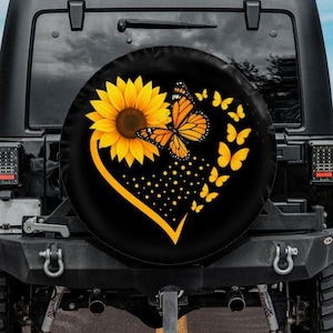 May include: Black spare tire cover featuring a yellow sunflower and orange butterfly design. The design includes a yellow heart shape and several yellow butterflies. The cover is mounted on the back of a black Jeep.
