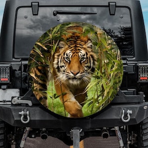 May include: A black Jeep with a spare tire cover featuring a tiger emerging from bamboo. The tiger has orange and black stripes, with green bamboo leaves surrounding it. The background shows a road and mountains.