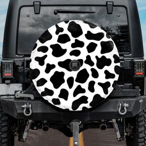 May include: A black Jeep with a spare tire cover featuring a white background and black cow spots. The vehicle is parked on a paved road with yellow lines, with a desert landscape and mountains in the background.