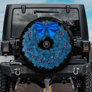 May include: A black Jeep SUV with a spare tire cover featuring a blue wreath design and a large blue bow. The wreath is decorated with small pink and blue ornaments. The vehicle is parked on a paved road with a mountain backdrop.