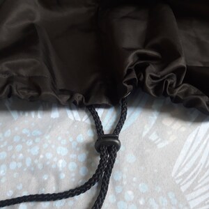 May include: Close-up of a black drawstring bag. The bag is made of a shiny, dark fabric. A black drawstring with a plastic adjuster is visible, along with a portion of the bag's opening.