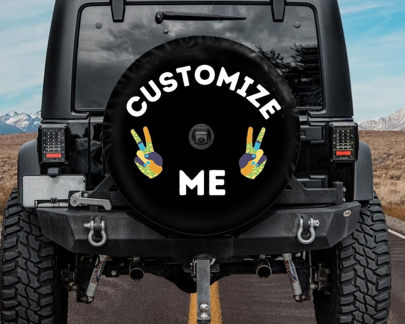 May include: Black spare tire cover with the words "CUSTOMIZE ME" in white. The cover features two peace sign hand gestures with colorful patterns. The cover is mounted on the back of a black Jeep.