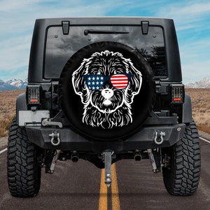 May include: A black spare tire cover with a white silhouette of a dog wearing sunglasses with red, white, and blue stars and stripes. The dog is looking directly at the camera.