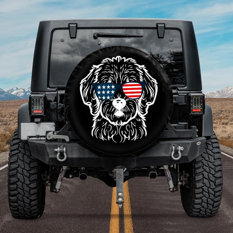 May include: A black spare tire cover with a white silhouette of a dog wearing sunglasses with red, white, and blue stars and stripes. The dog is looking directly at the camera.