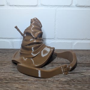 Könnte beinhalten: Ein brauner Sprechender Hut aus der Harry-Potter-Reihe. Der Hut ist aus einem weichen, biegsamen Material gefertigt und hat eine charakteristische, faltige Textur. Der Hut ist oben offen und hat einen Rand, der sich nach unten um die Seiten biegt. Der Hut liegt auf einer Holzoberfläche.