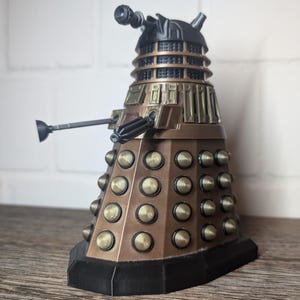 Könnte beinhalten: Eine goldene und schwarze Dalek-Figur mit einer schwarzen Waffe und einem schwarzen Sockel. Der Dalek hat einen kuppelförmigen Kopf mit einem schwarzen Auge und einer schwarzen Antenne.