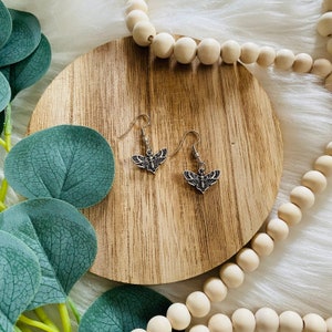 May include: A pair of silver dangle earrings with a detailed butterfly design. The earrings are on a wooden surface with a string of wooden beads.