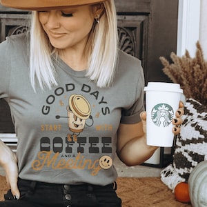 Puede incluir: Una mujer con sombrero marrón, camiseta gris y vaqueros negros, sosteniendo una taza de café Starbucks. La camiseta tiene el texto "Good Days Start With Coffee and a Meeting" con un gráfico de taza de café. La mujer está sentada en una alfombra estampada.