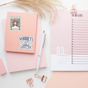 Puede incluir: Un cuaderno rosa con pegatinas que dicen "Mental Health Matters" y "Sobriety Social Club". Un bol&iacute;grafo blanco, una lista de tareas y clips de encuadernaci&oacute;n de oro rosa tambi&eacute;n est&aacute;n en la superficie rosa. Espigas de trigo secas en el fondo.