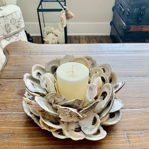May include: A white candle sits in the center of a circular arrangement of oyster shells. The shells are arranged in a flower-like pattern and are a light beige color.