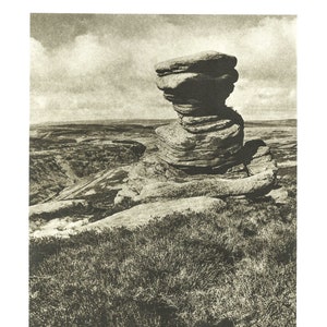 Puede incluir: Una fotografía en blanco y negro de una gran formación rocosa, que se asemeja a una cabeza, de pie en la cima de una colina cubierta de hierba. La formación rocosa está erosionada y tiene un aspecto estratificado. El fondo muestra un paisaje distante.