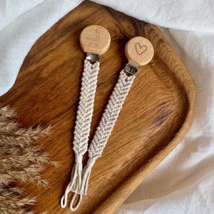 Puede incluir: Dos clips de chupete de macramé con tapas redondas de madera. Un clip presenta un grabado de jirafa, el otro un corazón. Los clips son beige y están sujetos a cierres metálicos. El fondo es una bandeja de madera y tela blanca.
