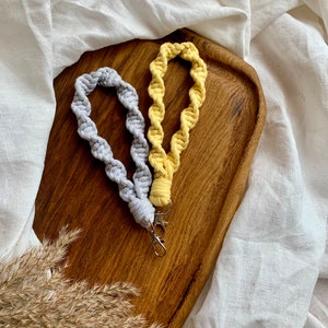 May include: Two handmade macrame keychains, one in grey and one in yellow, on a wooden surface.