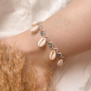 Puede incluir: Una tobillera de macram&eacute; beige adornada con peque&ntilde;as conchas de color crema y cuentas turquesas. La tobillera se lleva en un tobillo, mostrando la delicada artesan&iacute;a y la est&eacute;tica playera. Un accesorio elegante.