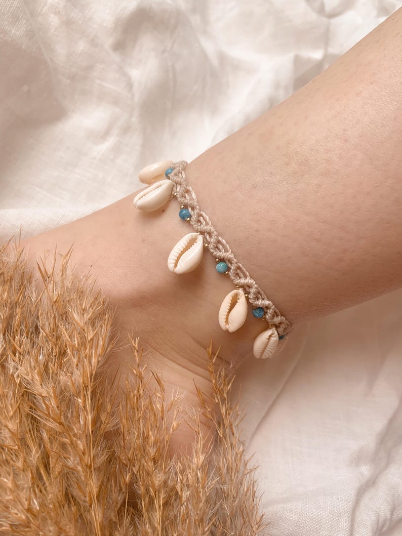 Puede incluir: Una tobillera de macram&eacute; beige adornada con peque&ntilde;as conchas de color crema y cuentas turquesas. La tobillera se lleva en un tobillo, mostrando la delicada artesan&iacute;a y la est&eacute;tica playera. Un accesorio elegante.