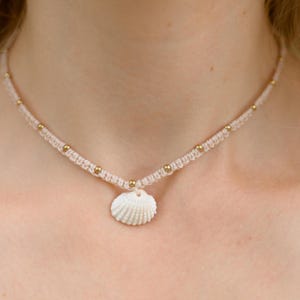 May include: A delicate white seashell necklace with a gold bead accent. The necklace is made of a thin, braided cord and features a small, white seashell pendant.
