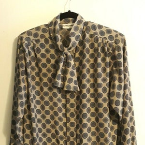 Puede incluir: Una blusa beige de manga larga con un patrón geométrico y una corbata de lazo. La blusa tiene un dobladillo con volantes y está hecha por Alfredo Durante.