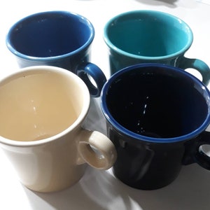 Könnte beinhalten: Vier Keramikbecher in Blau-, Türkis- und Cremetönen. Jeder Becher hat einen abgerundeten Henkel und eine leicht konische Form. Die Becher sind quadratisch angeordnet und zeigen ihre verschiedenen Farben und Designs.