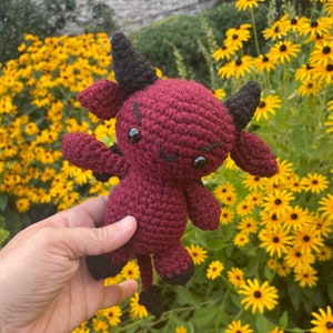 May include: A crocheted red devil plush toy with black horns and eyes. The toy is being held in a hand against a background of yellow flowers.