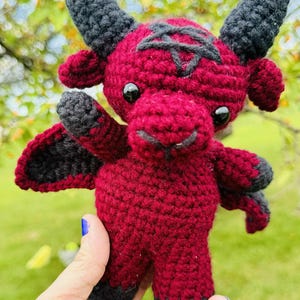 May include: A handmade crochet doll of a red baphomet with black horns, wings, and hooves. The doll features a pentagram on its head and black button eyes. The doll is held by a person.