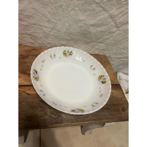 May include: White oval serving dish with a scalloped edge. The dish is decorated with small, colorful floral and house motifs around the rim. The dish is set on a wooden surface.