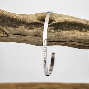 Könnte beinhalten: Ein silbernes Armreif-Armband mit den Koordinaten "73.3228°N - 41.7211°W" eingraviert.