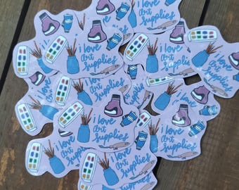 Puede incluir: Una colección de pegatinas troqueladas sobre un fondo morado claro, cada una con ilustraciones de una paleta de acuarelas, un tarro azul con pinceles, un tubo de pintura y un tintero, con el texto I love art supplies escrito en azul.