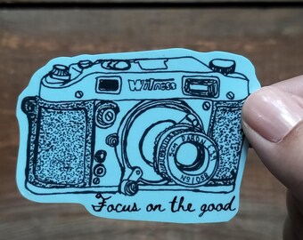 Puede incluir: Una pegatina azul claro con una ilustración en negro de una cámara vintage. La cámara tiene la palabra "Witness" en la parte superior y la frase "Focus on the good" en la parte inferior. La pegatina la sostiene una persona.