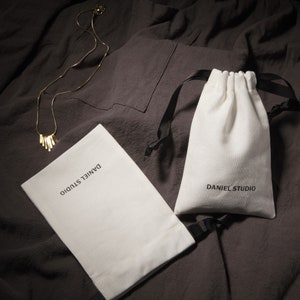 May include: A gold-colored necklace with a bar pendant rests on a dark gray fabric. Two white drawstring pouches with the text "DANIEL STUDIO" are also present. One pouch is open, and the other is closed.