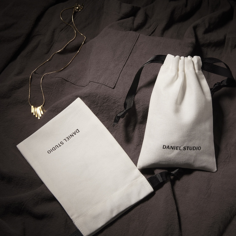 May include: A gold-colored necklace with a bar pendant rests on a dark gray fabric. Two white drawstring pouches with the text "DANIEL STUDIO" are also present. One pouch is open, and the other is closed.