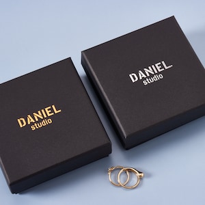 May include: Two black boxes with the text "DANIEL studio" printed in gold on the top. The boxes are on a light blue surface. A pair of gold hoop earrings are in front of the boxes.
