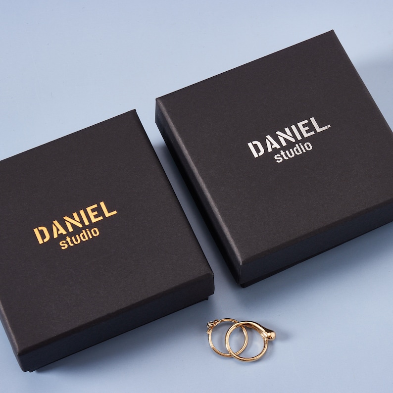 May include: Two black boxes with the text "DANIEL studio" printed in gold on the top. The boxes are on a light blue surface. A pair of gold hoop earrings are in front of the boxes.